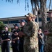 A group of U.S. Air Force personnel and civilian firefighters are gathered outdoors on what appears to be a military installation, likely during a ceremony or formal event. The individuals are standing at attention or applauding, wearing a mix of OCP (Operational Camouflage Pattern) uniforms and dark firefighter duty uniforms, several bearing 'FIRE' patches. The setting includes palm trees and a multi-story building consistent with a military base in a warm-climate region.