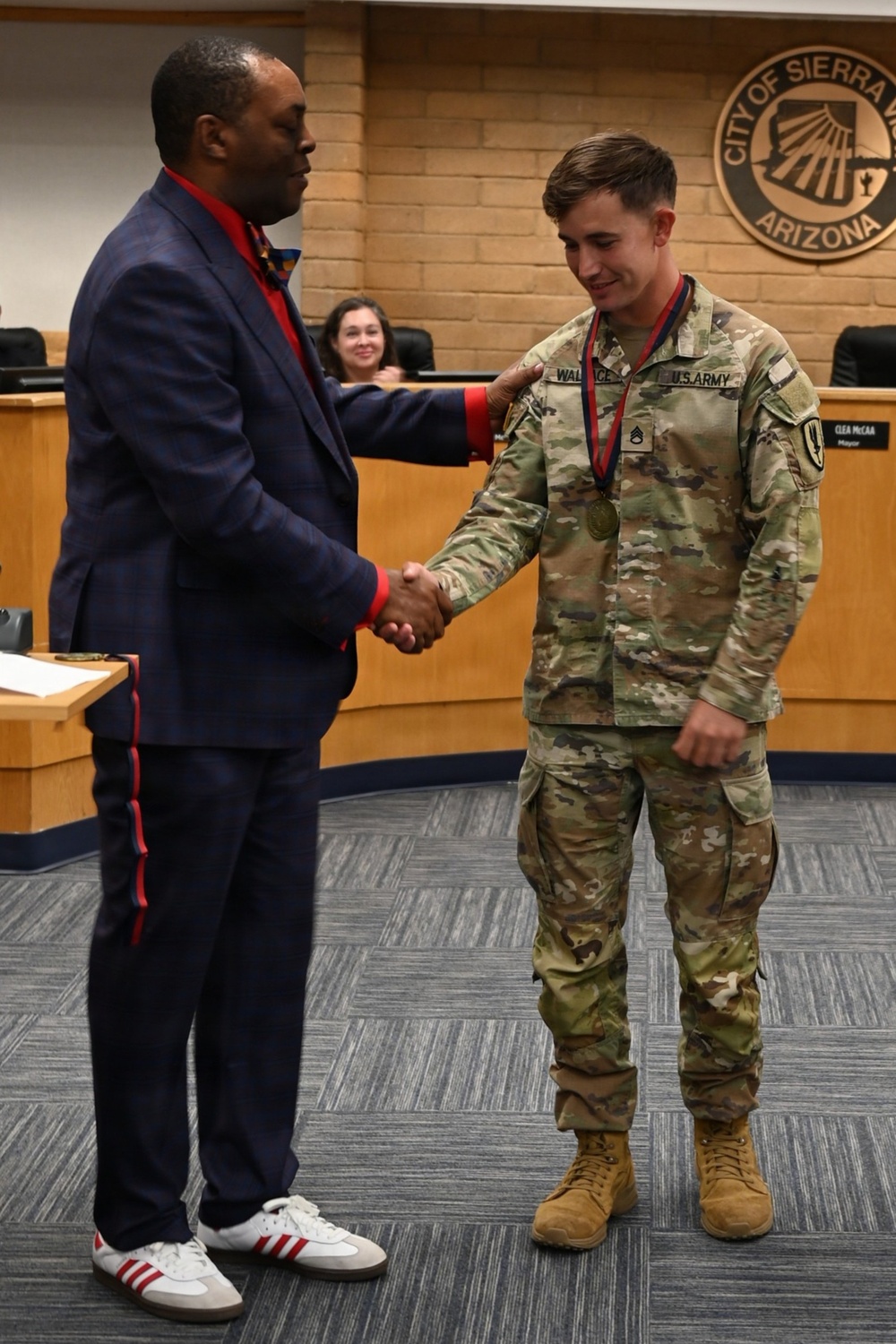 A U.S. Army soldier wearing OCP (Operational Camouflage Pattern) uniform with the name tape 'WALLACE' is receiving a medal during a ceremony at the City of Sierra Vista, Arizona city council chambers. A civilian man in a navy blue suit is shaking the soldier's hand and placing a hand on his shoulder in a congratulatory gesture. A woman is visible smiling in the background near the council dais.