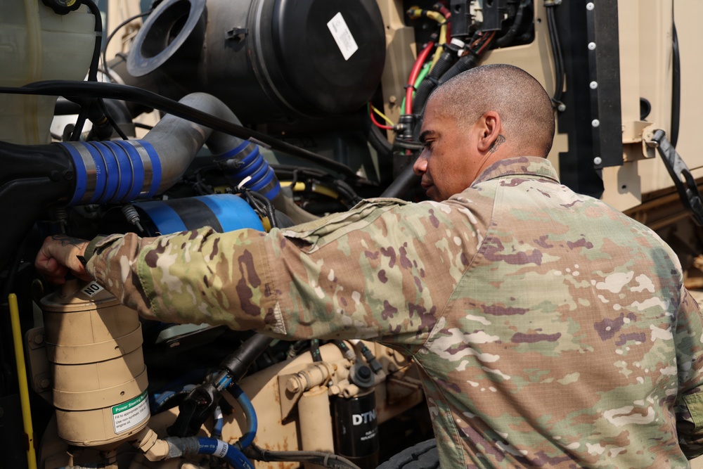 A U.S. military service member in OCP (Operational Camouflage Pattern) uniform is performing maintenance on a military vehicle engine compartment. The soldier is closely inspecting or adjusting components within the engine bay, which contains various hoses, filters, and mechanical parts. The setting appears to be an outdoor military environment with tan/desert-colored equipment.