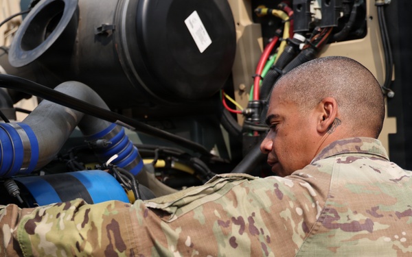 A U.S. military service member in OCP (Operational Camouflage Pattern) uniform is performing maintenance on a military vehicle engine compartment. The soldier is closely inspecting or adjusting components within the engine bay, which contains various hoses, filters, and mechanical parts. The setting appears to be an outdoor military environment with tan/desert-colored equipment.