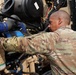 A U.S. military service member in OCP (Operational Camouflage Pattern) uniform is performing maintenance on a military vehicle engine compartment. The soldier is closely inspecting or adjusting components within the engine bay, which contains various hoses, filters, and mechanical parts. The setting appears to be an outdoor military environment with tan/desert-colored equipment.