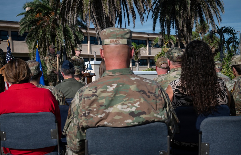 An outdoor military ceremony is taking place, likely at a U.S. Air Force installation, with personnel in OCP (Operational Camouflage Pattern) uniforms seated as an audience facing a speaker at a podium. A ceremonial bell is visible on a table near the front, and military flags including the U.S. flag are present. The setting features palm trees and a multi-story building in the background, suggesting a warm-climate base.