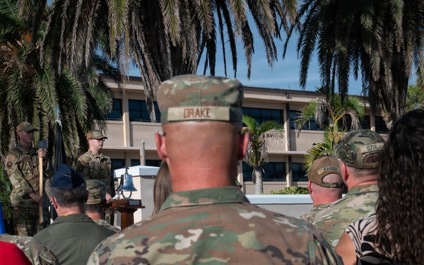 An outdoor military ceremony is taking place, likely at a U.S. Air Force installation, with personnel in OCP (Operational Camouflage Pattern) uniforms seated as an audience facing a speaker at a podium. A ceremonial bell is visible on a table near the front, and military flags including the U.S. flag are present. The setting features palm trees and a multi-story building in the background, suggesting a warm-climate base.
