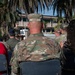 An outdoor military ceremony is taking place, likely at a U.S. Air Force installation, with personnel in OCP (Operational Camouflage Pattern) uniforms seated as an audience facing a speaker at a podium. A ceremonial bell is visible on a table near the front, and military flags including the U.S. flag are present. The setting features palm trees and a multi-story building in the background, suggesting a warm-climate base.