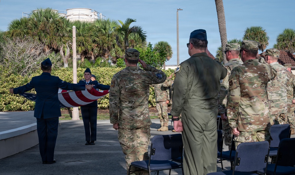 A military ceremony is taking place outdoors at what appears to be a U.S. Air Force installation, likely in a subtropical location given the palm trees and vegetation. Two honor guard members in dress blue uniforms are folding an American flag while officers in flight suits and soldiers in OCP (Operational Camouflage Pattern) uniforms render salutes. The setting includes a paved plaza area with folding chairs arranged for an audience.