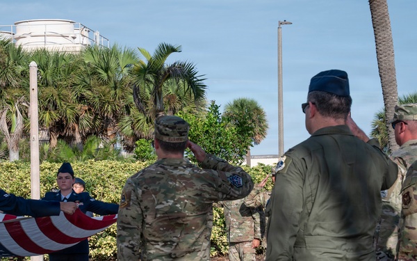 A military ceremony is taking place outdoors at what appears to be a U.S. Air Force installation, likely in a subtropical location given the palm trees and vegetation. Two honor guard members in dress blue uniforms are folding an American flag while officers in flight suits and soldiers in OCP (Operational Camouflage Pattern) uniforms render salutes. The setting includes a paved plaza area with folding chairs arranged for an audience.