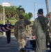 A military ceremony is taking place outdoors at what appears to be a U.S. Air Force installation, likely in a subtropical location given the palm trees and vegetation. Two honor guard members in dress blue uniforms are folding an American flag while officers in flight suits and soldiers in OCP (Operational Camouflage Pattern) uniforms render salutes. The setting includes a paved plaza area with folding chairs arranged for an audience.