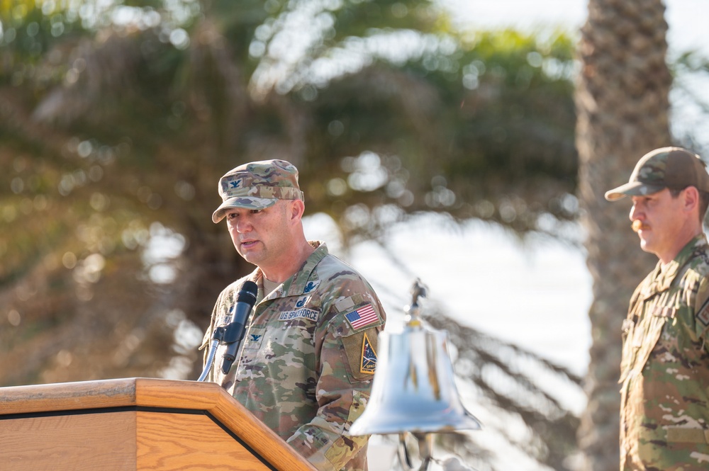 A U.S. Space Force military officer in OCP (Operational Camouflage Pattern) uniform is speaking at a wooden podium during what appears to be an outdoor ceremony. A silver bell is visible on the podium, and a second uniformed service member stands at attention in the background. The setting includes palm trees, suggesting a warm-climate military installation.