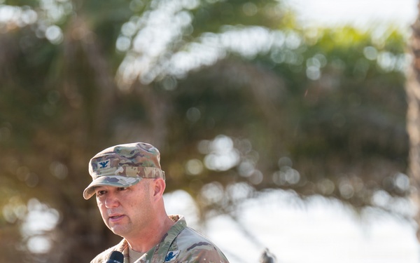 A U.S. Space Force military officer in OCP (Operational Camouflage Pattern) uniform is speaking at a wooden podium during what appears to be an outdoor ceremony. A silver bell is visible on the podium, and a second uniformed service member stands at attention in the background. The setting includes palm trees, suggesting a warm-climate military installation.