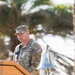 A U.S. Space Force military officer in OCP (Operational Camouflage Pattern) uniform is speaking at a wooden podium during what appears to be an outdoor ceremony. A silver bell is visible on the podium, and a second uniformed service member stands at attention in the background. The setting includes palm trees, suggesting a warm-climate military installation.