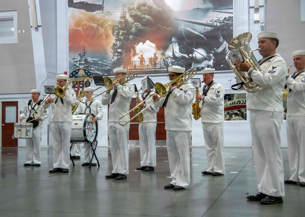 A U.S. Navy military band performs indoors, with musicians dressed in white Navy dress uniforms. The ensemble includes brass and percussion instruments, performing in front of a large mural depicting naval warfare history. A circular emblem visible in the background references 'America's Navy 250,' suggesting a commemorative anniversary event.