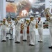 A U.S. Navy military band performs indoors, with musicians dressed in white Navy dress uniforms. The ensemble includes brass and percussion instruments, performing in front of a large mural depicting naval warfare history. A circular emblem visible in the background references 'America's Navy 250,' suggesting a commemorative anniversary event.