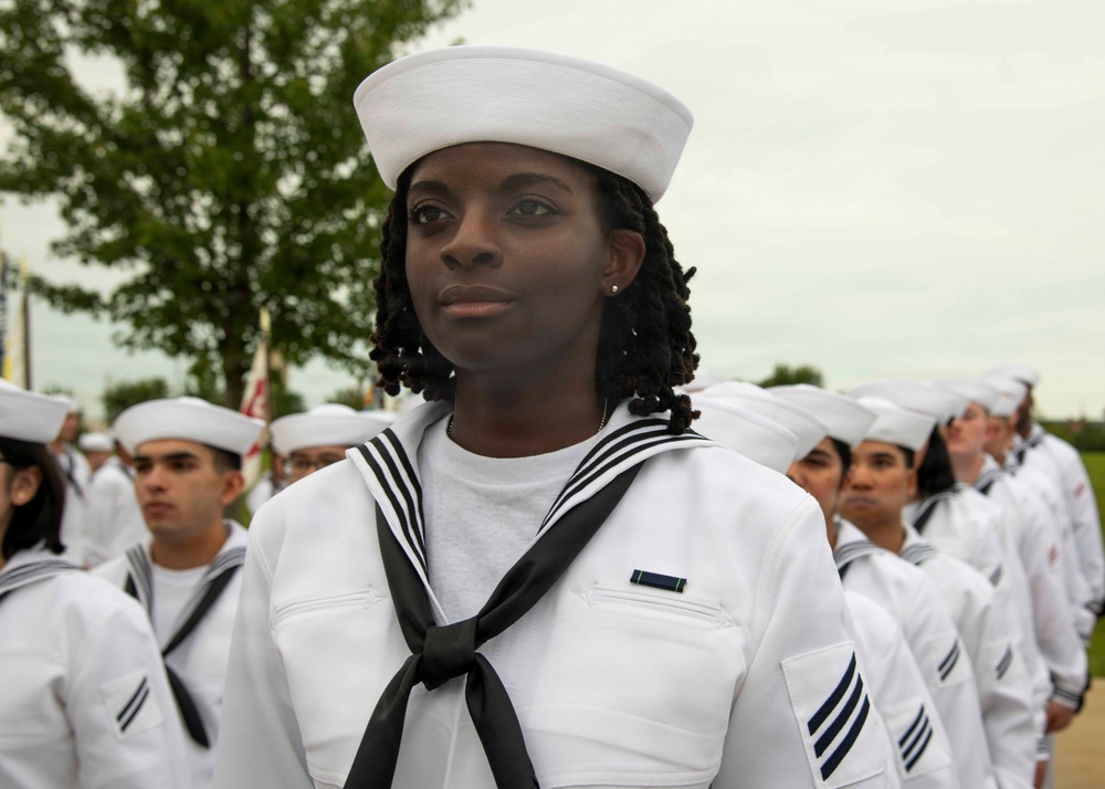A group of U.S. Navy personnel in dress white uniforms are standing in formation, likely during a ceremony or parade. The foreground subject is a female sailor wearing a white dixie cup hat, black neckerchief, and displaying rating chevrons and a service ribbon on her uniform. The background shows additional sailors in similar uniforms along with trees and an overcast sky.