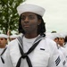A group of U.S. Navy personnel in dress white uniforms are standing in formation, likely during a ceremony or parade. The foreground subject is a female sailor wearing a white dixie cup hat, black neckerchief, and displaying rating chevrons and a service ribbon on her uniform. The background shows additional sailors in similar uniforms along with trees and an overcast sky.