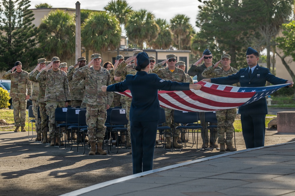 A military flag ceremony is taking place outdoors at what appears to be a U.S. military installation, likely in a tropical or subtropical location based on the palm trees in the background. Two service members in dress blue Air Force uniforms are unfolding or folding the American flag while a formation of military personnel in camouflage uniforms render a salute. The ceremony appears formal and organized, consistent with a retreat or reveille flag ceremony.