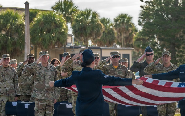 A military flag ceremony is taking place outdoors at what appears to be a U.S. military installation, likely in a tropical or subtropical location based on the palm trees in the background. Two service members in dress blue Air Force uniforms are unfolding or folding the American flag while a formation of military personnel in camouflage uniforms render a salute. The ceremony appears formal and organized, consistent with a retreat or reveille flag ceremony.