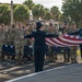A military flag ceremony is taking place outdoors at what appears to be a U.S. military installation, likely in a tropical or subtropical location based on the palm trees in the background. Two service members in dress blue Air Force uniforms are unfolding or folding the American flag while a formation of military personnel in camouflage uniforms render a salute. The ceremony appears formal and organized, consistent with a retreat or reveille flag ceremony.