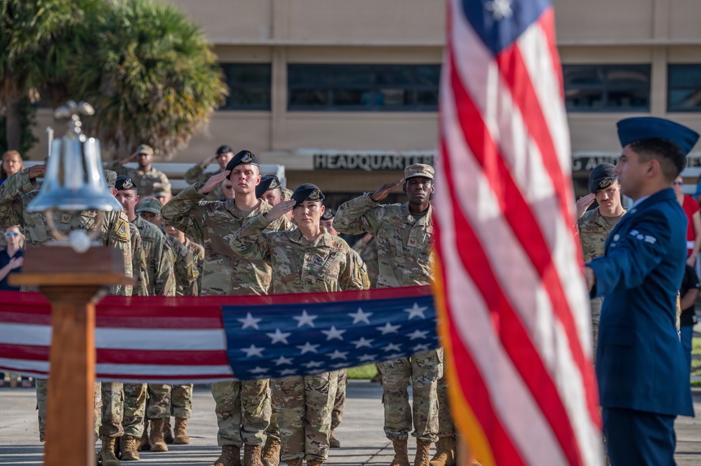 A military ceremony is taking place outdoors in front of a building marked 'Headquarters.' Personnel in camouflage uniforms and black berets are rendering salutes alongside an officer in dress blue uniform, with American flags prominently displayed and a ceremonial bell on a wooden podium visible in the foreground.
