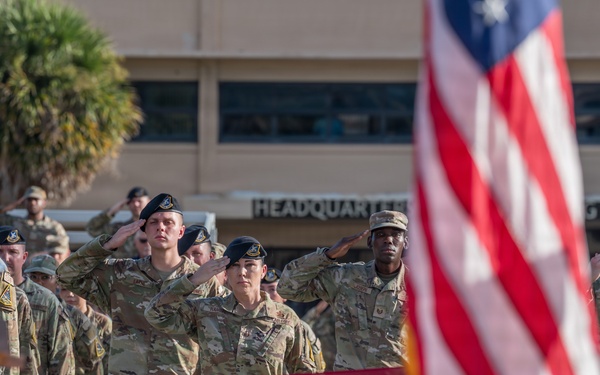 A military ceremony is taking place outdoors in front of a building marked 'Headquarters.' Personnel in camouflage uniforms and black berets are rendering salutes alongside an officer in dress blue uniform, with American flags prominently displayed and a ceremonial bell on a wooden podium visible in the foreground.