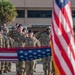 A military ceremony is taking place outdoors in front of a building marked 'Headquarters.' Personnel in camouflage uniforms and black berets are rendering salutes alongside an officer in dress blue uniform, with American flags prominently displayed and a ceremonial bell on a wooden podium visible in the foreground.