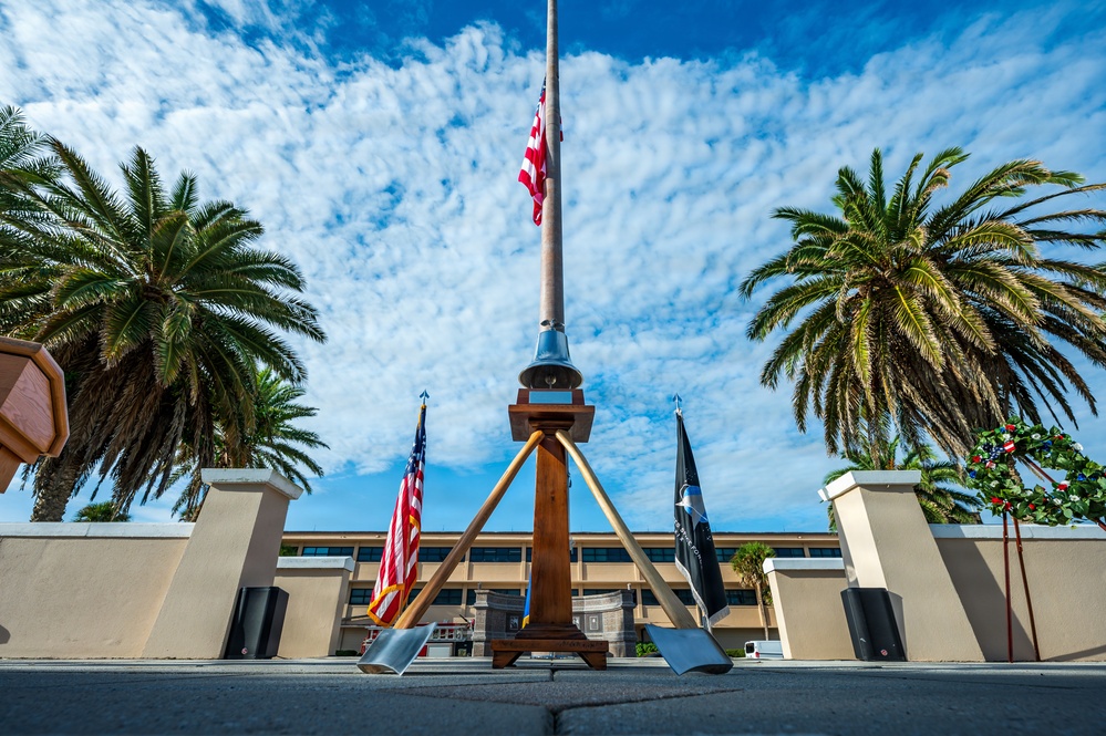 A low-angle photograph of a formal outdoor memorial or ceremonial area featuring a tall flagpole flying the American flag at half-staff, flanked by palm trees and a wooden tripod structure with a bell. Two flags — an American flag and what appears to be a POW/MIA flag — stand on either side of the central structure. The setting appears to be a military installation or government facility, with a multi-story building visible in the background.