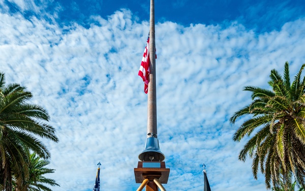 A low-angle photograph of a formal outdoor memorial or ceremonial area featuring a tall flagpole flying the American flag at half-staff, flanked by palm trees and a wooden tripod structure with a bell. Two flags — an American flag and what appears to be a POW/MIA flag — stand on either side of the central structure. The setting appears to be a military installation or government facility, with a multi-story building visible in the background.