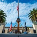 A low-angle photograph of a formal outdoor memorial or ceremonial area featuring a tall flagpole flying the American flag at half-staff, flanked by palm trees and a wooden tripod structure with a bell. Two flags — an American flag and what appears to be a POW/MIA flag — stand on either side of the central structure. The setting appears to be a military installation or government facility, with a multi-story building visible in the background.