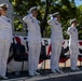 Five U.S. Navy officers in white dress uniforms are standing at salute during a formal military ceremony. The event appears to be held outdoors on a ship's deck or platform decorated with American flag bunting. The setting and attire suggest a commissioning, change of command, or similar official naval ceremony.