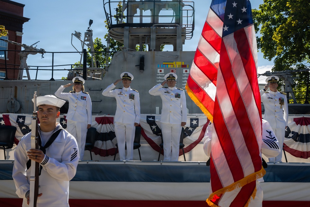 A formal U.S. Navy military ceremony is taking place in front of a naval vessel. Four officers in dress white uniforms stand on an elevated platform rendering salutes, while a sailor in the foreground carries the U.S. flag with gold fringe. The setting is decorated with patriotic bunting in red, white, and blue.