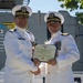 Two U.S. Navy officers in white dress uniforms are shaking hands while jointly holding a certificate. The certificate is visibly titled 'Meritorious Service Medal' issued by the United States of America. The ceremony appears to be taking place outdoors, likely aboard a naval vessel or installation.