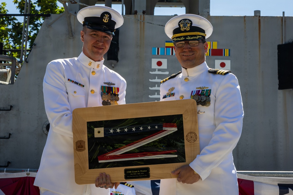 Two U.S. Navy officers in white dress uniforms are posing together while holding a framed shadow box containing a folded American flag. The plaque on the frame reads 'CDR Grant A. Wanier' with dates suggesting a change of command or retirement ceremony. The ceremony appears to be taking place outdoors on or near a naval vessel.