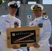 Two U.S. Navy officers in white dress uniforms are posing together while holding a framed shadow box containing a folded American flag. The plaque on the frame reads 'CDR Grant A. Wanier' with dates suggesting a change of command or retirement ceremony. The ceremony appears to be taking place outdoors on or near a naval vessel.
