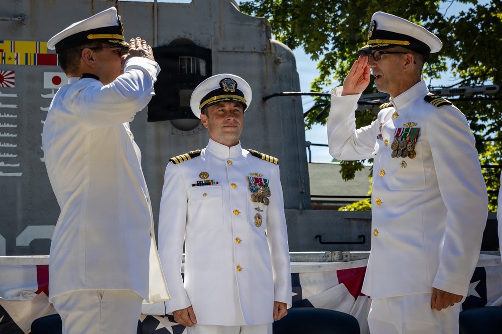 Three U.S. Navy officers in white dress uniforms are participating in a formal military ceremony, likely a change of command. Two officers are exchanging salutes while a third stands at attention between them. The ceremony appears to be taking place outdoors near a naval vessel, with signal flags and an American flag visible in the background.