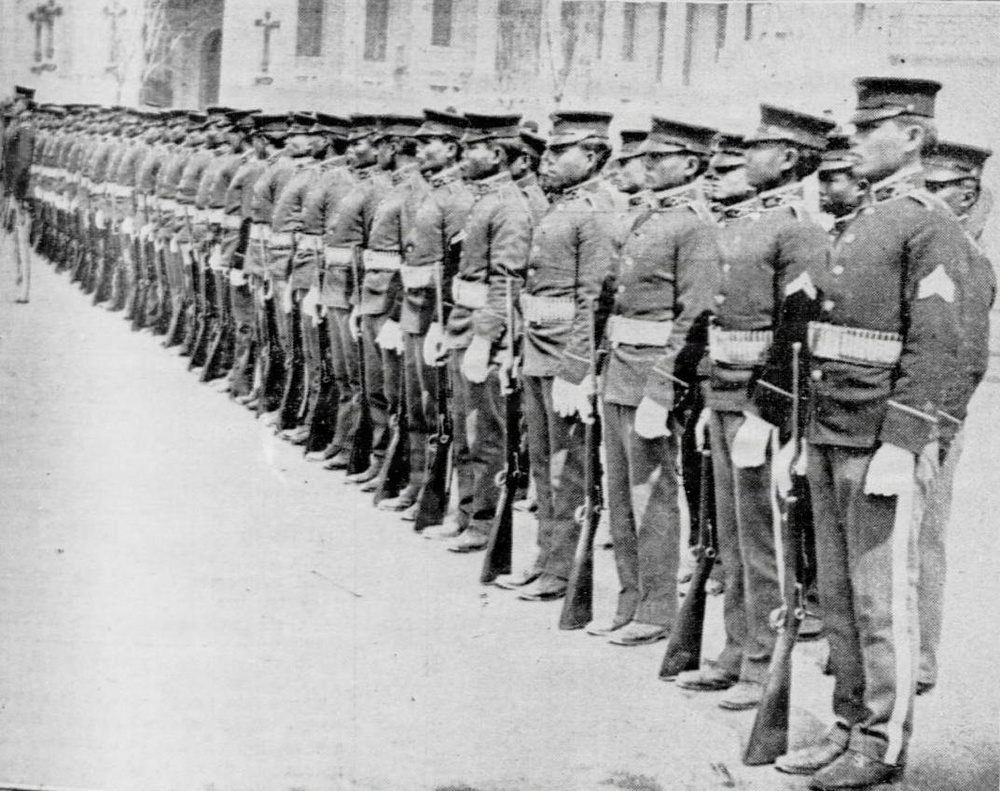 A black-and-white archival photograph showing a large formation of uniformed soldiers standing at attention in what appears to be a military parade or inspection. The soldiers are arranged in a long diagonal line extending into the background, all wearing matching military uniforms with caps and carrying rifles. A large building is visible in the background, suggesting an urban or institutional setting.