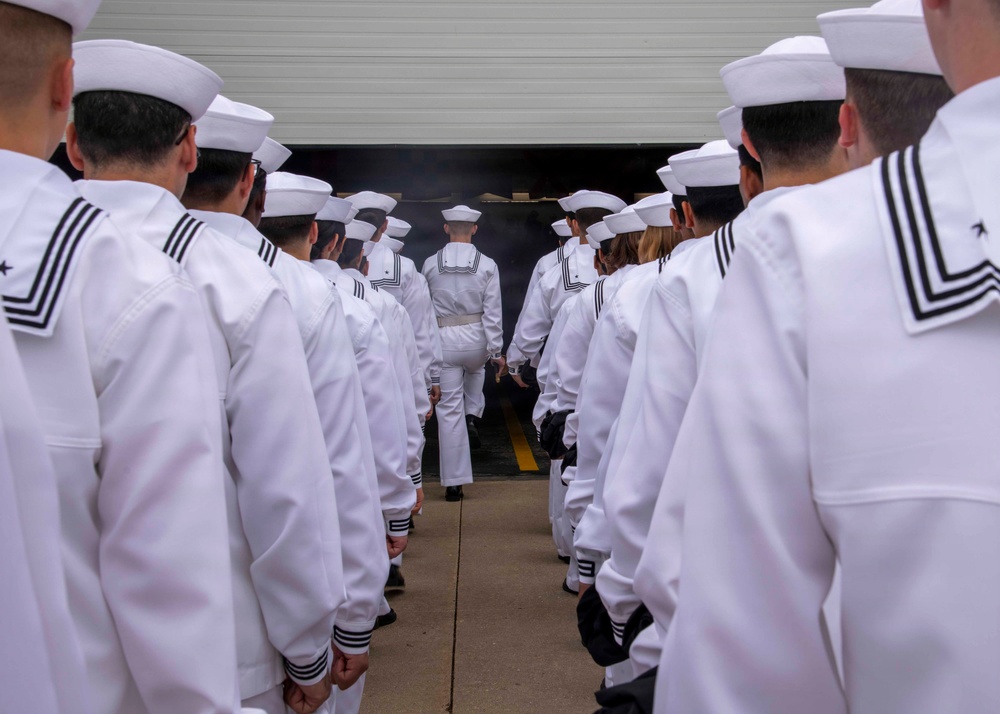 A formation of U.S. Navy sailors dressed in white dress uniforms and white Dixie cup covers are standing in two parallel lines facing a large open hangar or building entrance. The perspective is from behind the formation, looking toward the entrance where smoke or haze is visible. The scene appears to be a formal military ceremony, commissioning event, or official parade.
