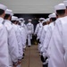 A formation of U.S. Navy sailors dressed in white dress uniforms and white Dixie cup covers are standing in two parallel lines facing a large open hangar or building entrance. The perspective is from behind the formation, looking toward the entrance where smoke or haze is visible. The scene appears to be a formal military ceremony, commissioning event, or official parade.