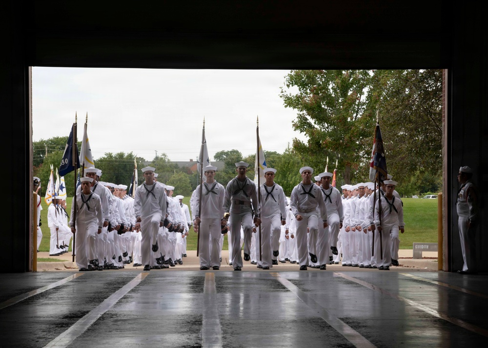 A large formation of U.S. Navy personnel dressed in white dress uniforms are marching in a ceremonial parade, entering through a large open bay or hangar doorway. Color guard members carry multiple flags at the front of the formation. The scene appears to be a formal military graduation or pass-in-review ceremony conducted on a wet, overcast day.