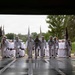 A large formation of U.S. Navy personnel dressed in white dress uniforms are marching in a ceremonial parade, entering through a large open bay or hangar doorway. Color guard members carry multiple flags at the front of the formation. The scene appears to be a formal military graduation or pass-in-review ceremony conducted on a wet, overcast day.