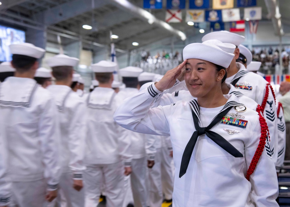 A U.S. Navy sailor in dress white uniform is rendering a salute during what appears to be a formal military ceremony or graduation event held indoors. The foreground sailor is a young woman wearing multiple service ribbons, a warfare pin, and a red aiguillette, indicating a position of distinction. Multiple sailors in matching dress whites are visible in the background, along with military flags and pennants hanging from the ceiling.