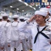 A U.S. Navy sailor in dress white uniform is rendering a salute during what appears to be a formal military ceremony or graduation event held indoors. The foreground sailor is a young woman wearing multiple service ribbons, a warfare pin, and a red aiguillette, indicating a position of distinction. Multiple sailors in matching dress whites are visible in the background, along with military flags and pennants hanging from the ceiling.