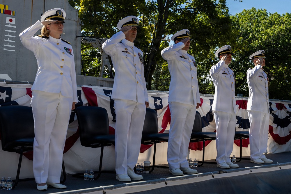 Five U.S. Navy officers in white dress uniforms are standing at salute on a raised platform during a formal military ceremony. The setting is decorated with American flag bunting, suggesting a commissioning, change of command, or similar official naval event. The officers include both male and female personnel displaying various service ribbons and insignia.