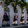 Five U.S. Navy officers in white dress uniforms are standing at salute on a raised platform during a formal military ceremony. The setting is decorated with American flag bunting, suggesting a commissioning, change of command, or similar official naval event. The officers include both male and female personnel displaying various service ribbons and insignia.