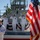 A formal U.S. Navy military ceremony is taking place in front of a naval vessel. Four officers in dress white uniforms stand on an elevated platform rendering salutes, while a sailor in the foreground carries the U.S. flag with gold fringe. The setting is decorated with patriotic bunting in red, white, and blue.
