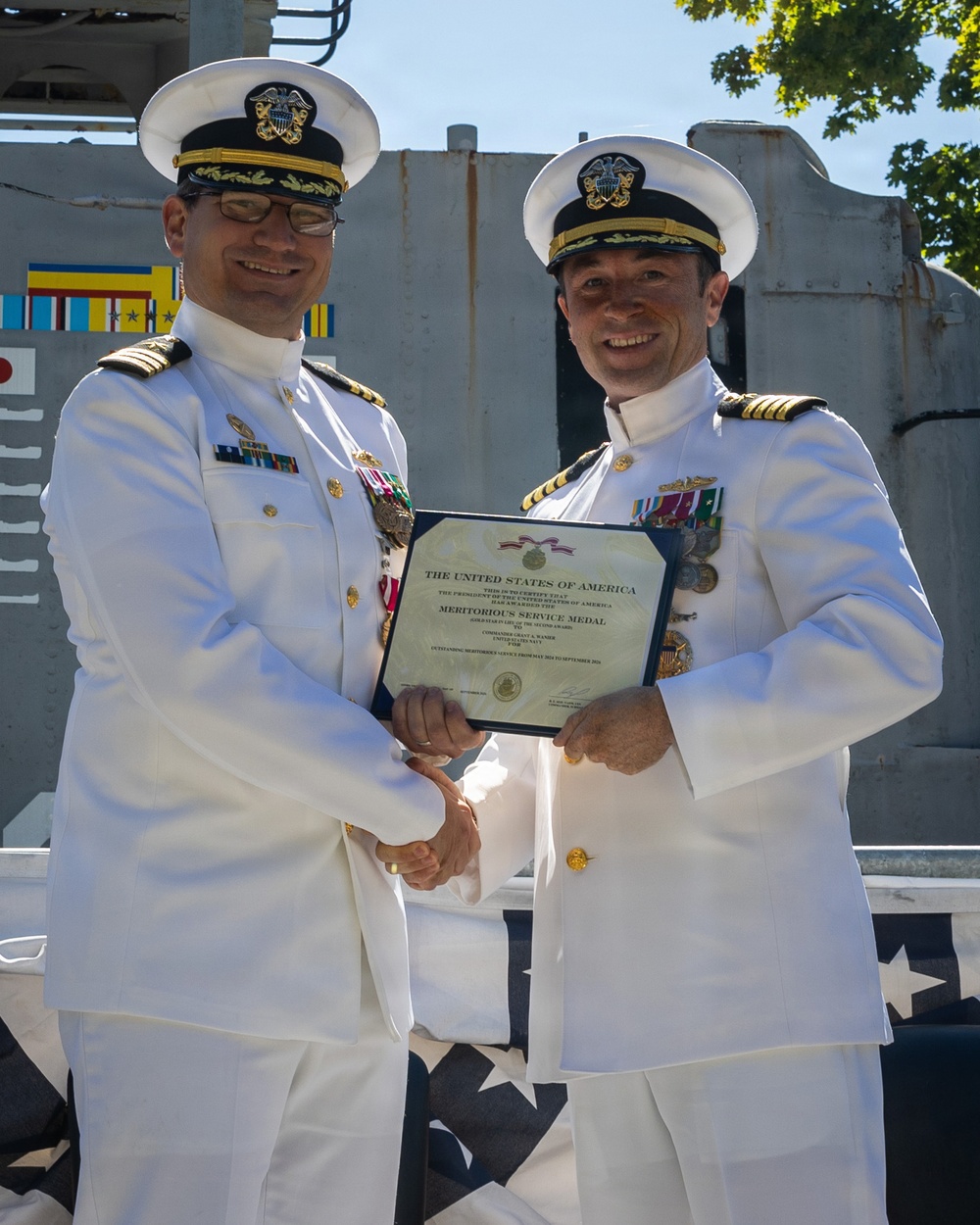 Two U.S. Navy officers in white dress uniforms are shaking hands while jointly holding a certificate. The certificate is visibly titled 'Meritorious Service Medal' issued by the United States of America. The ceremony appears to be taking place outdoors, likely aboard a naval vessel or installation.