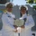 Two U.S. Navy officers in white dress uniforms are shaking hands while jointly holding a certificate. The certificate is visibly titled 'Meritorious Service Medal' issued by the United States of America. The ceremony appears to be taking place outdoors, likely aboard a naval vessel or installation.
