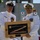 Two U.S. Navy officers in white dress uniforms are posing together while holding a framed shadow box containing a folded American flag. The plaque on the frame reads 'CDR Grant A. Wanier' with dates suggesting a change of command or retirement ceremony. The ceremony appears to be taking place outdoors on or near a naval vessel.
