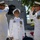 Three U.S. Navy officers in white dress uniforms are participating in a formal military ceremony, likely a change of command. Two officers on the left and right are exchanging salutes while a third officer stands at attention in the center. The ceremony appears to be taking place outdoors near a naval vessel, with signal flags and an American flag visible in the background.