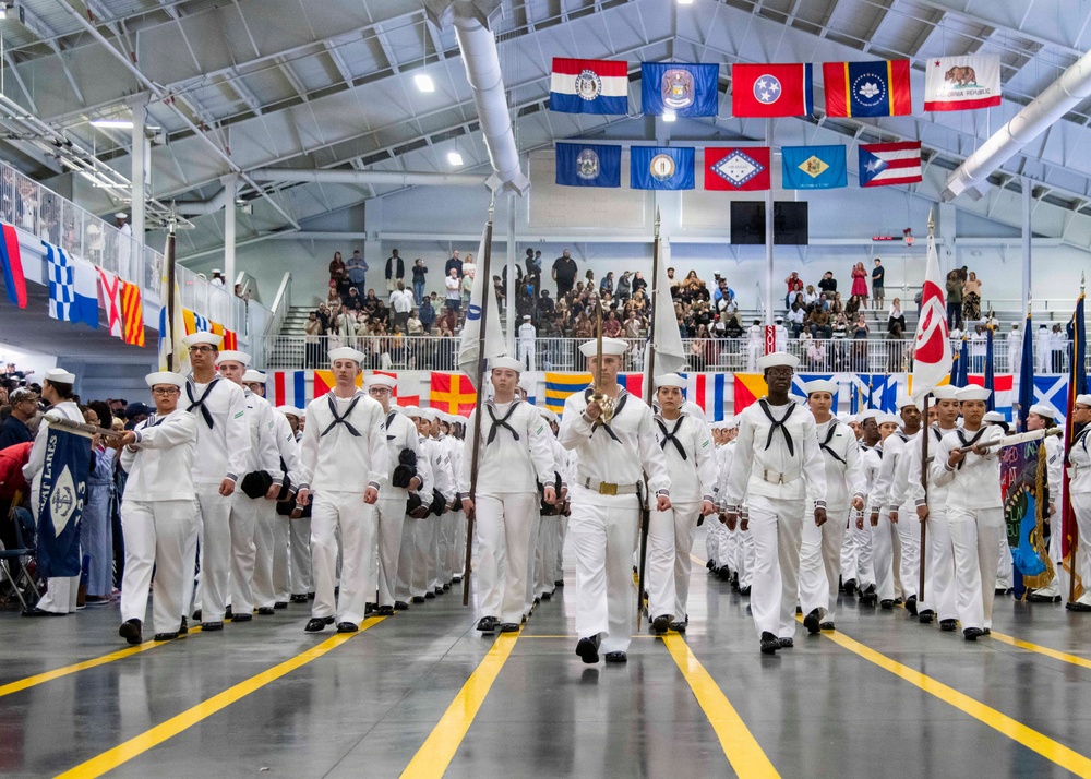 A large formation of U.S. Navy recruits in dress white uniforms marches in a ceremonial parade inside a large indoor drill hall. The event appears to be a Navy boot camp graduation ceremony, likely at Naval Station Great Lakes in Illinois, as suggested by a partially visible 'Great Lakes' banner on the left. Spectators fill the bleachers in the background, and numerous U.S. state flags and naval signal flags are displayed throughout the venue.