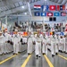 A large formation of U.S. Navy recruits in dress white uniforms marches in a ceremonial parade inside a large indoor drill hall. The event appears to be a Navy boot camp graduation ceremony, likely at Naval Station Great Lakes in Illinois, as suggested by a partially visible 'Great Lakes' banner on the left. Spectators fill the bleachers in the background, and numerous U.S. state flags and naval signal flags are displayed throughout the venue.