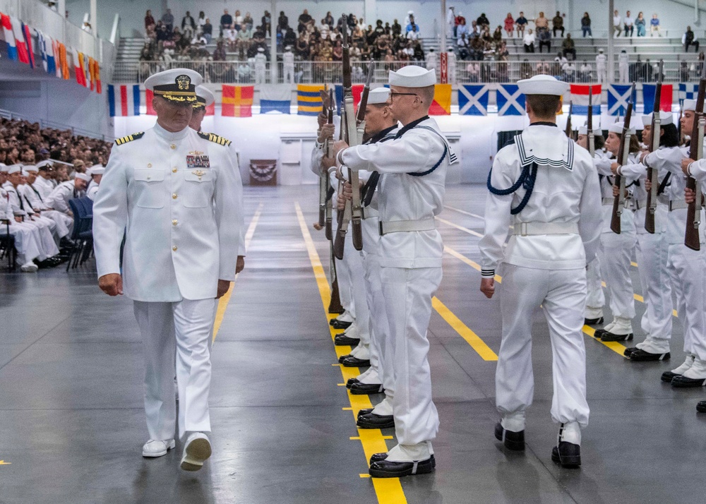 A formal U.S. Navy military ceremony taking place in a large indoor arena or hangar. A senior officer in dress whites with gold insignia walks past a rifle honor guard also dressed in white uniforms. A large audience of spectators and seated sailors fills the background, with international and naval signal flags decorating the walls.